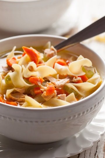 Bowl of turkey noodle soup with a spoon resting on a white scalloped plate.