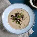 Bowl of creamy chestnut mushroom soup in a blue bowl and garnished with sauteed mushrooms and fresh herbs.