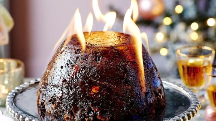 A traditional Christmas pudding, lovingly made on Stir-Up Sunday, is set alight and served on a decorative cake stand, with festive decorations and a Christmas tree in the background.