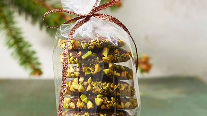 Stack of chocolate cookies coated with chopped pistachios, packaged in a clear plastic bag tied with a ribbon&mdash;perfect as Christmas food gifts&mdash;with two cookies beside the bag on a green surface.
