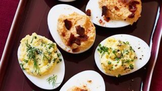 A rectangular plate of deviled eggs, topped with chopped herbs and crumbled bacon, is arranged neatly against a dark red background—perfect for your 12 Days of Christmas Recipes collection.