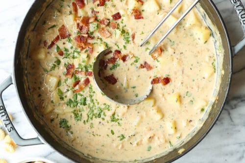 A pot of creamy chowder with potatoes and herbs, topped with chopped bacon-perfect for Christmas Eve recipes. Surrounding the pot are a bowl of oyster crackers, measuring spoons, and a bowl of bacon pieces.