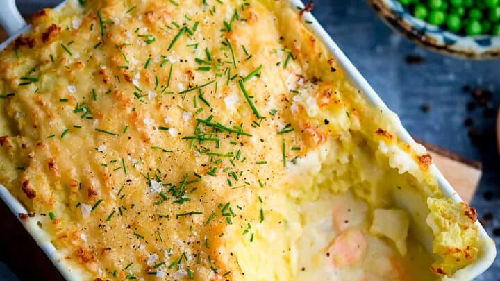 A rectangular dish of baked fish pie topped with mashed potatoes and herbs, with a portion served out—perfect for cozy Christmas Eve recipes; bowls of peas and corn are in the background.
