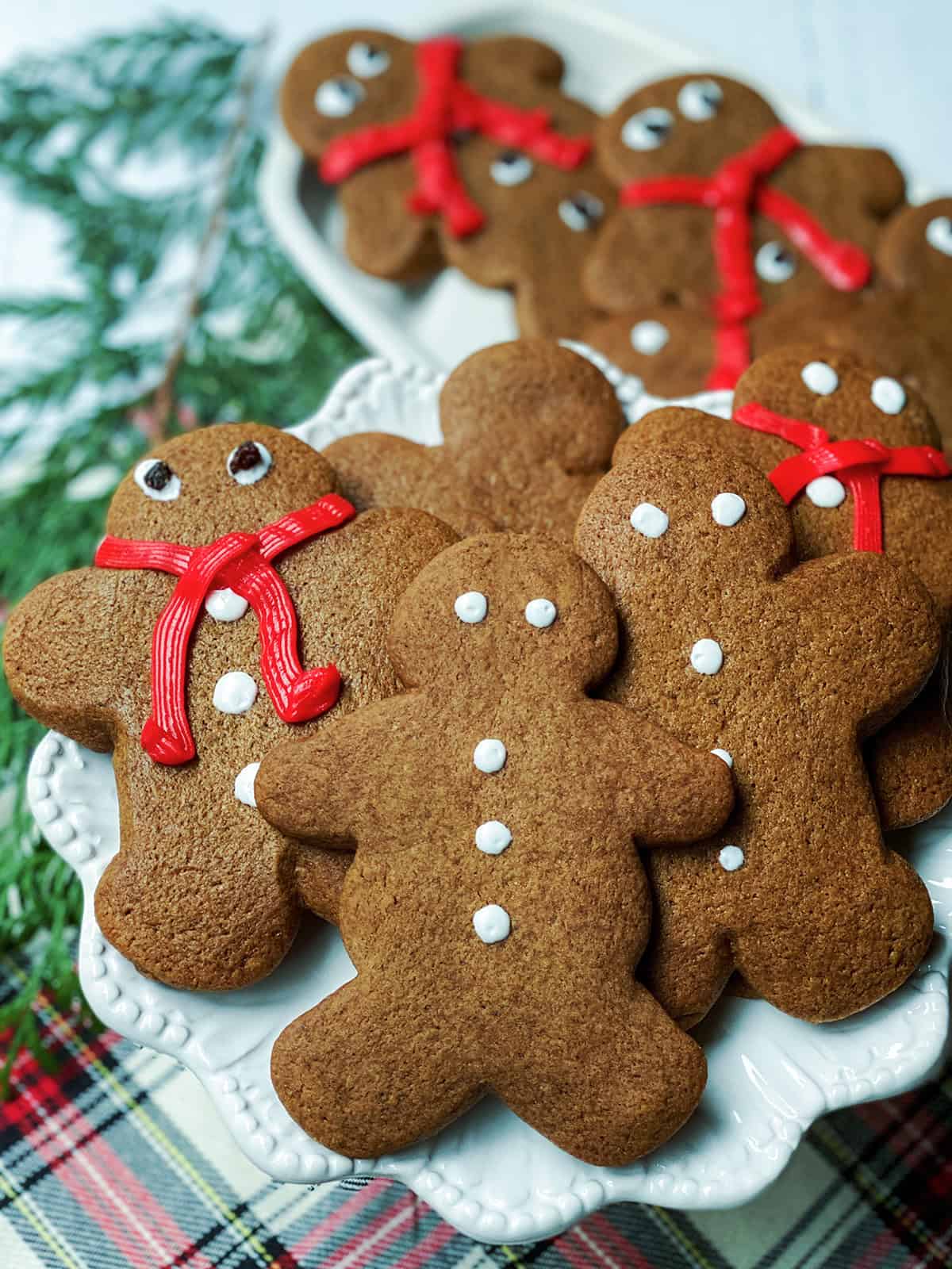 A plate of festive gingerbread cookies decorated with white and red icing, shaped like gingerbread people, sits on a patterned tablecloth with evergreen branches nearby.