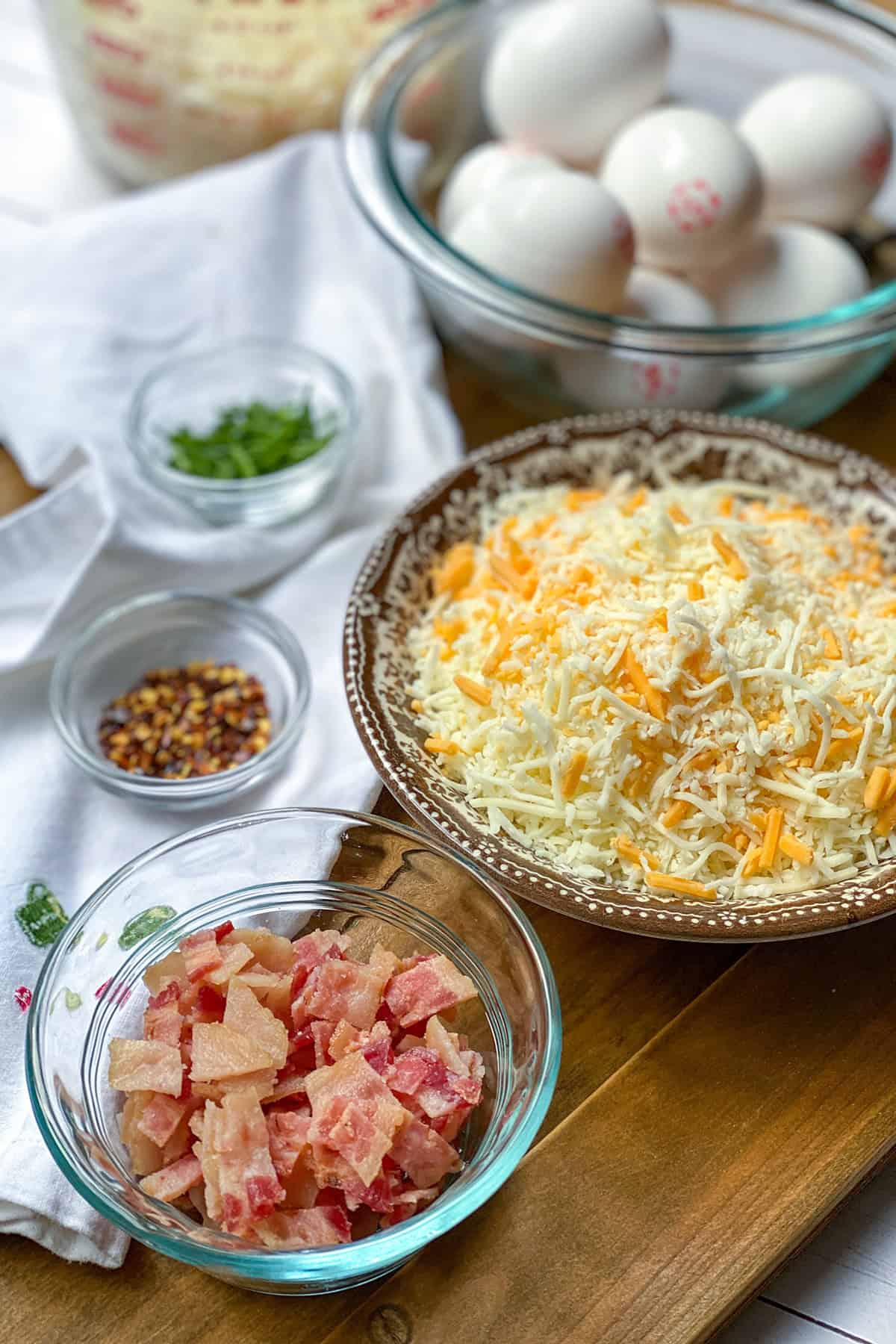 Bowls of shredded cheese, chopped cooked bacon, eggs, chopped chives, and red pepper flakes arranged on a wooden surface-perfect ingredients for a delicious hashbrown breakfast casserole.