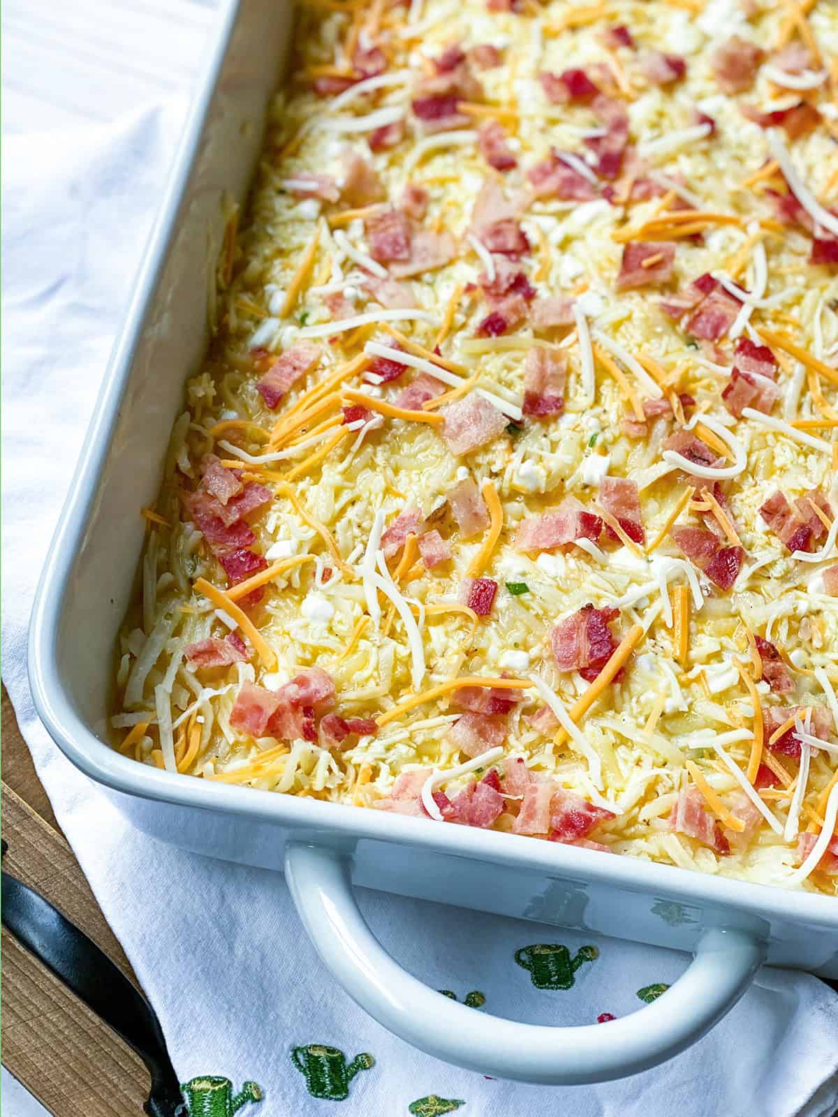 A close-up of a hashbrown breakfast casserole in a dish, filled with a baked mixture topped with melted shredded cheese and chopped bacon.
