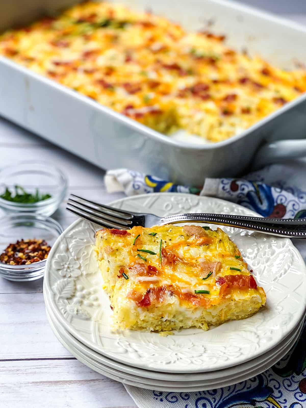 A slice of hashbrown breakfast casserole with cheese, ham, and chives on a plate with a fork, with the casserole dish and seasoning bowls in the background.