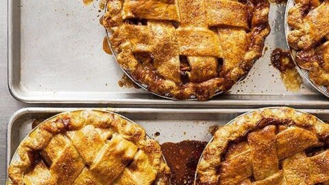 Several golden brown lattice-topped pies are arranged on a metal baking sheet, their filling and juices spilling over the edges—perfect for sharing as part of your 12 Days of Christmas Recipes celebration.