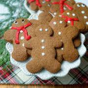 A plate of festive gingerbread cookies shaped like people, decorated with white icing and red icing scarves, rests on a plaid cloth next to fresh evergreen branches.