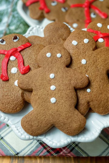 A plate of festive gingerbread cookies shaped like people, decorated with white icing and red icing scarves, rests on a plaid cloth next to fresh evergreen branches.