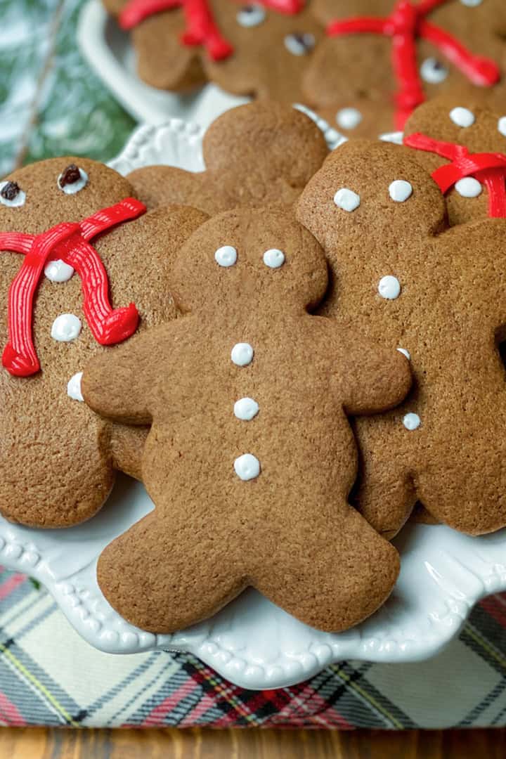 A plate of festive gingerbread cookies shaped like people, decorated with white icing and red icing scarves, rests on a plaid cloth next to fresh evergreen branches.