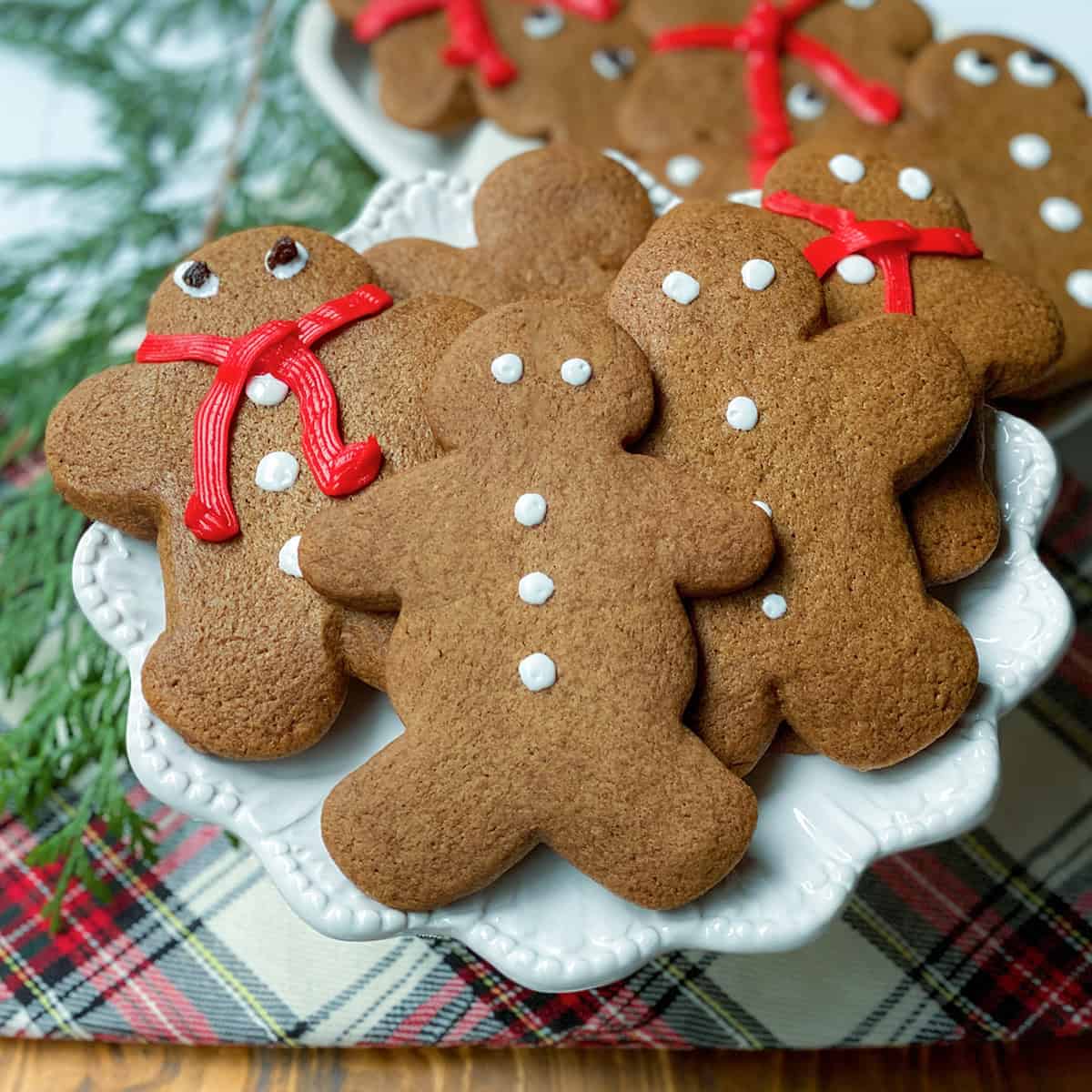 A plate of festive gingerbread cookies shaped like people, decorated with white icing and red icing scarves, rests on a plaid cloth next to fresh evergreen branches.