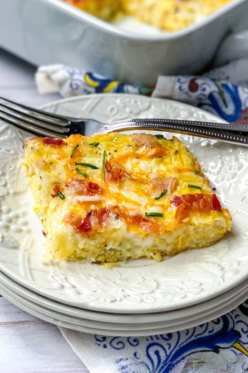 A slice of cheesy hashbrown breakfast casserole with fresh herbs sits on a white plate, a fork beside it and the casserole dish in the background.