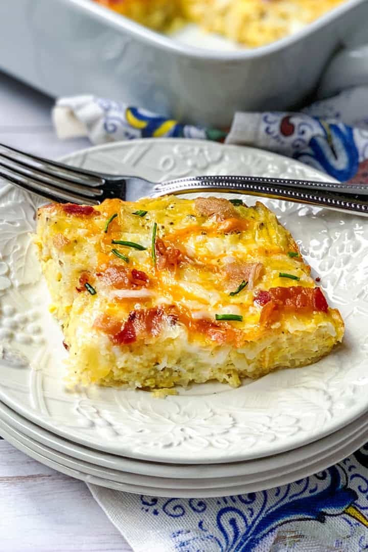 A slice of cheesy hashbrown breakfast casserole with fresh herbs sits on a white plate, a fork beside it and the casserole dish in the background.