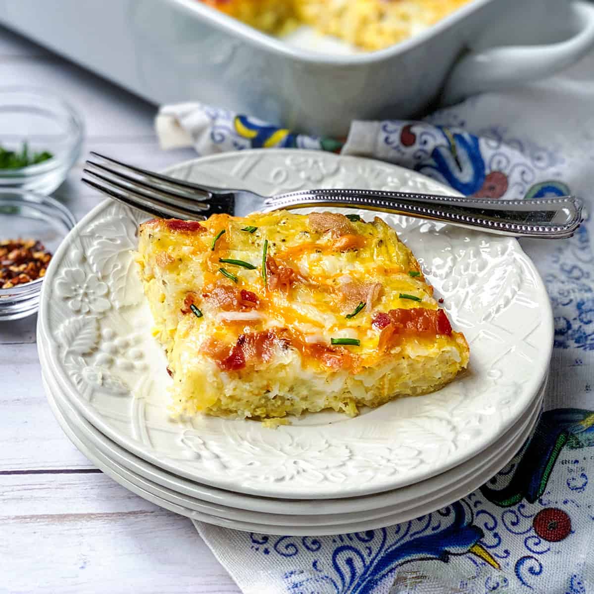 A slice of cheesy hashbrown breakfast casserole with fresh herbs sits on a white plate, a fork beside it and the casserole dish in the background.