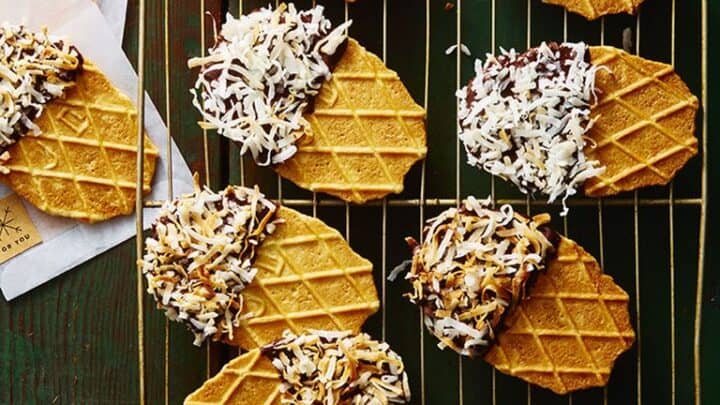 Waffle cookies on a cooling rack, some half-dipped in chocolate and sprinkled with shredded coconut, make delightful christmas food gifts on a rustic dark wood surface.