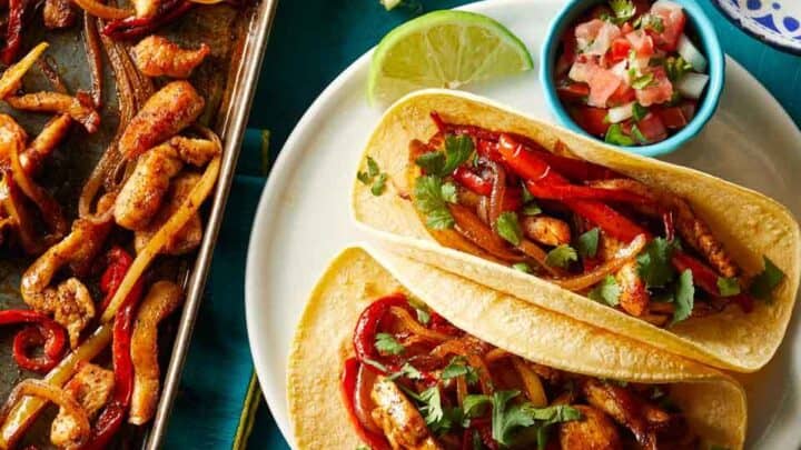 Two chicken fajita tacos with grilled peppers and onions on a white plate, garnished with cilantro, served with lime wedge and salsa—this easy Healthy Dump Dinners recipe sits next to a pan of fajita filling.