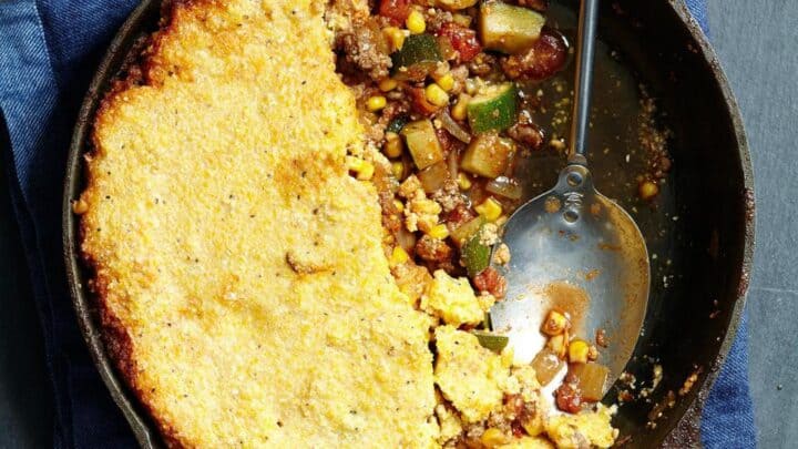 A cast iron skillet with a partially served casserole topped with cornbread, revealing a filling of ground meat, vegetables, and corn—an inviting example of Healthy Dump Dinners; a serving spoon rests inside.
