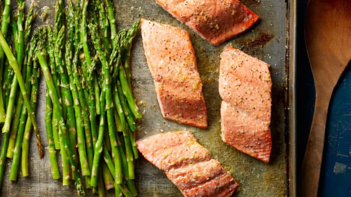 Baked salmon fillets and asparagus spears arranged on a metal sheet pan next to a wooden spatula, set against a dark blue surface—a perfect example of Healthy Dump Dinners.