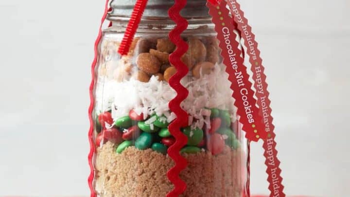 A mason jar filled with layered baking ingredients&mdash;flour, brown sugar, chocolate chips, and candies&mdash;decorated with a red ribbon and holiday tag on a red surface, makes delightful Christmas food gifts.