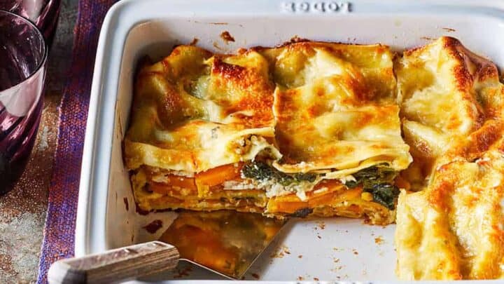 A square baking dish containing a partially served lasagna with visible layers of pasta, cheese, vegetables, and sauce—perfect for Christmas Week Dinners; a serving spatula and two empty glasses are nearby.