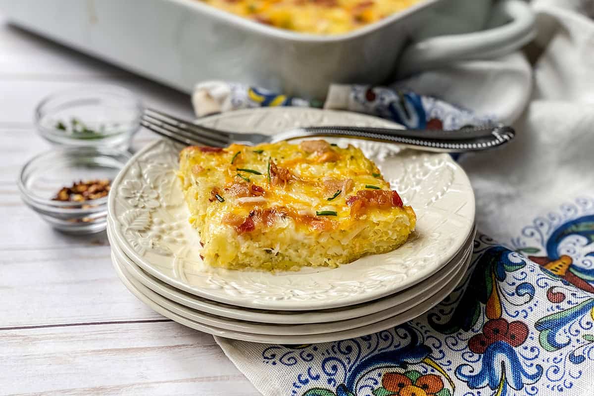 A square slice of hashbrown breakfast casserole with cheese and herbs on a stacked plate, accompanied by a fork and casserole dish in the background.