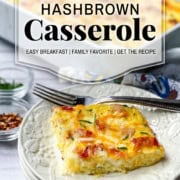 A slice of hashbrown breakfast casserole on a white plate with a fork, garnished with herbs, with a baking dish of casserole in the background.