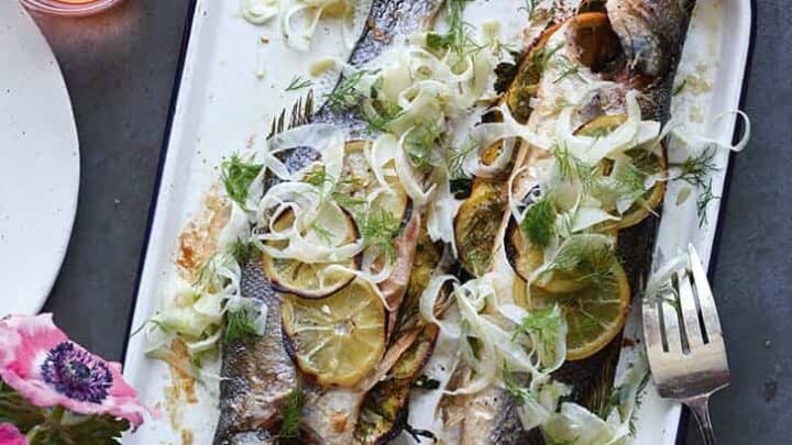Two whole baked fish garnished with lemon slices, fresh herbs, and fennel are served on a white tray—an elegant choice for Christmas Eve recipes, perfect for a festive table alongside drinks and a fork.