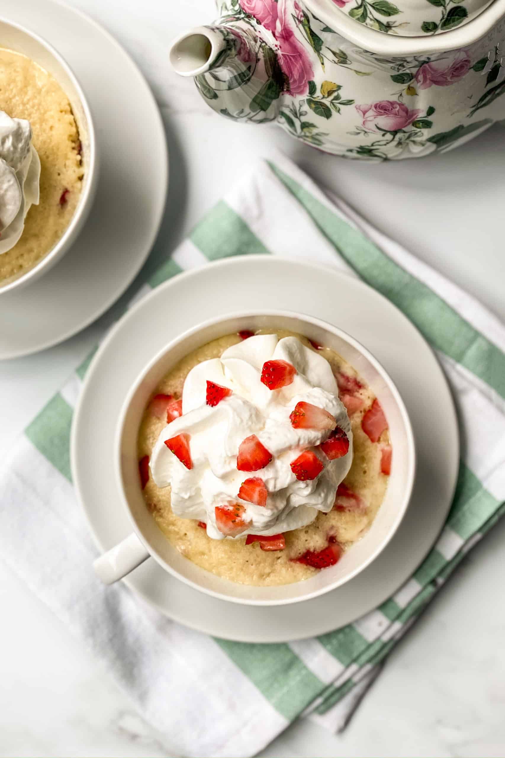A strawberry mug cake topped with whipped cream and fresh strawberry pieces sits on a white and green napkin beside a floral teapot.