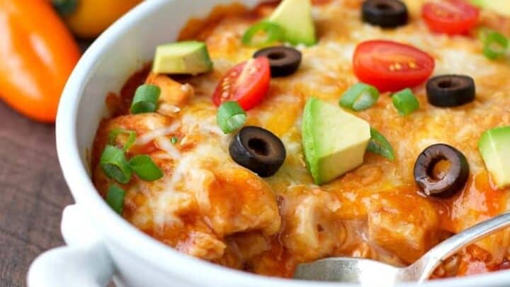 A white casserole dish filled with a baked cheesy Mexican-style dish, perfect for Healthy 5 Ingredient Dinners, topped with avocado, cherry tomatoes, black olives, and green onions. A spoon rests inside the dish.