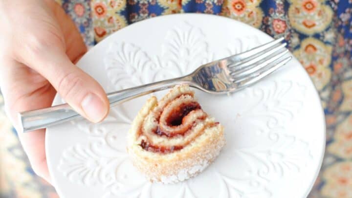 A hand holds a white plate with a single slice of jelly roll cake and a fork, perfect for fika, against the backdrop of a floral patterned dress.