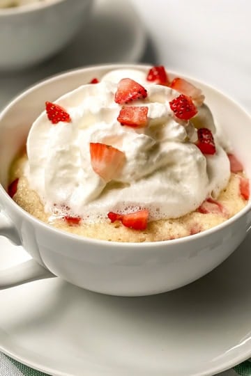 A white mug holds a delicious strawberry mug cake topped with whipped cream and chopped strawberries, placed on a saucer.