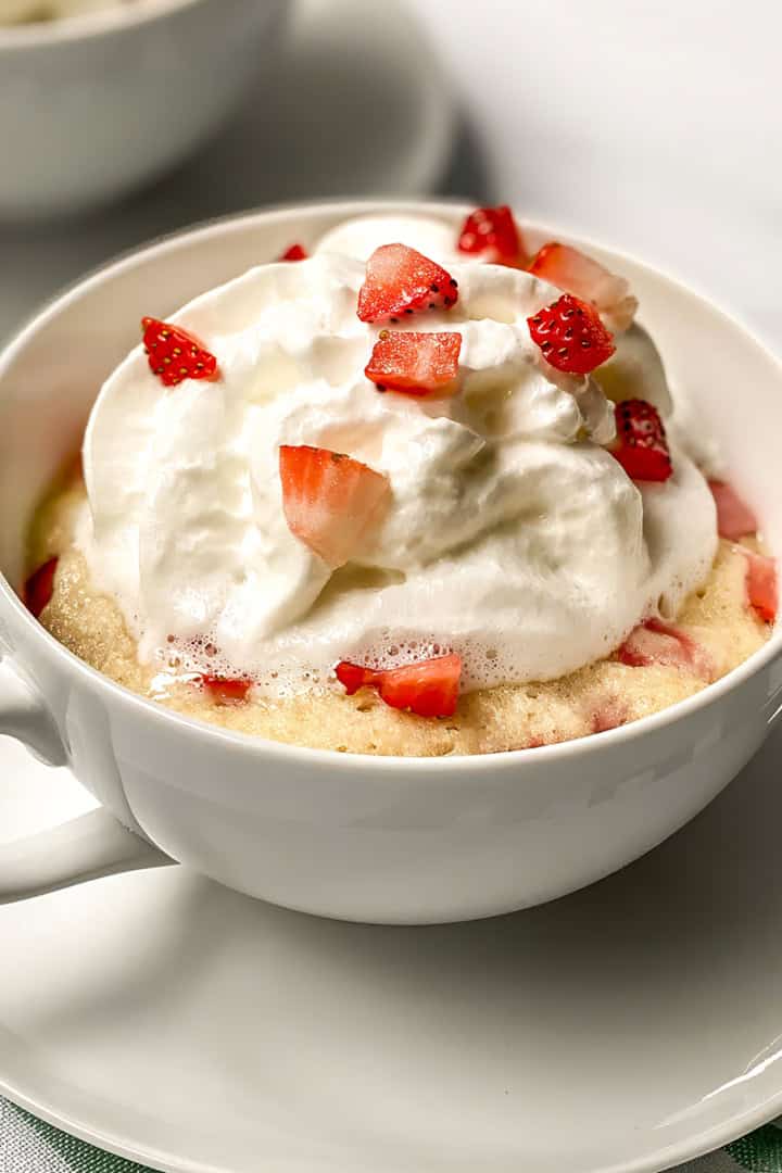A white mug holds a delicious strawberry mug cake topped with whipped cream and chopped strawberries, placed on a saucer.