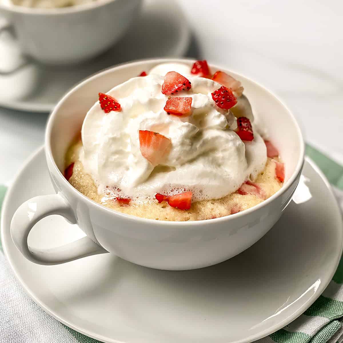 A white mug holds a delicious strawberry mug cake topped with whipped cream and chopped strawberries, placed on a saucer.