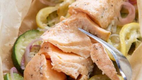 A hand holds a parchment packet with cooked salmon, sliced zucchini, lemon, and onions—an easy Healthy 5 Ingredient Dinners idea—while a fork gently flakes the fish.