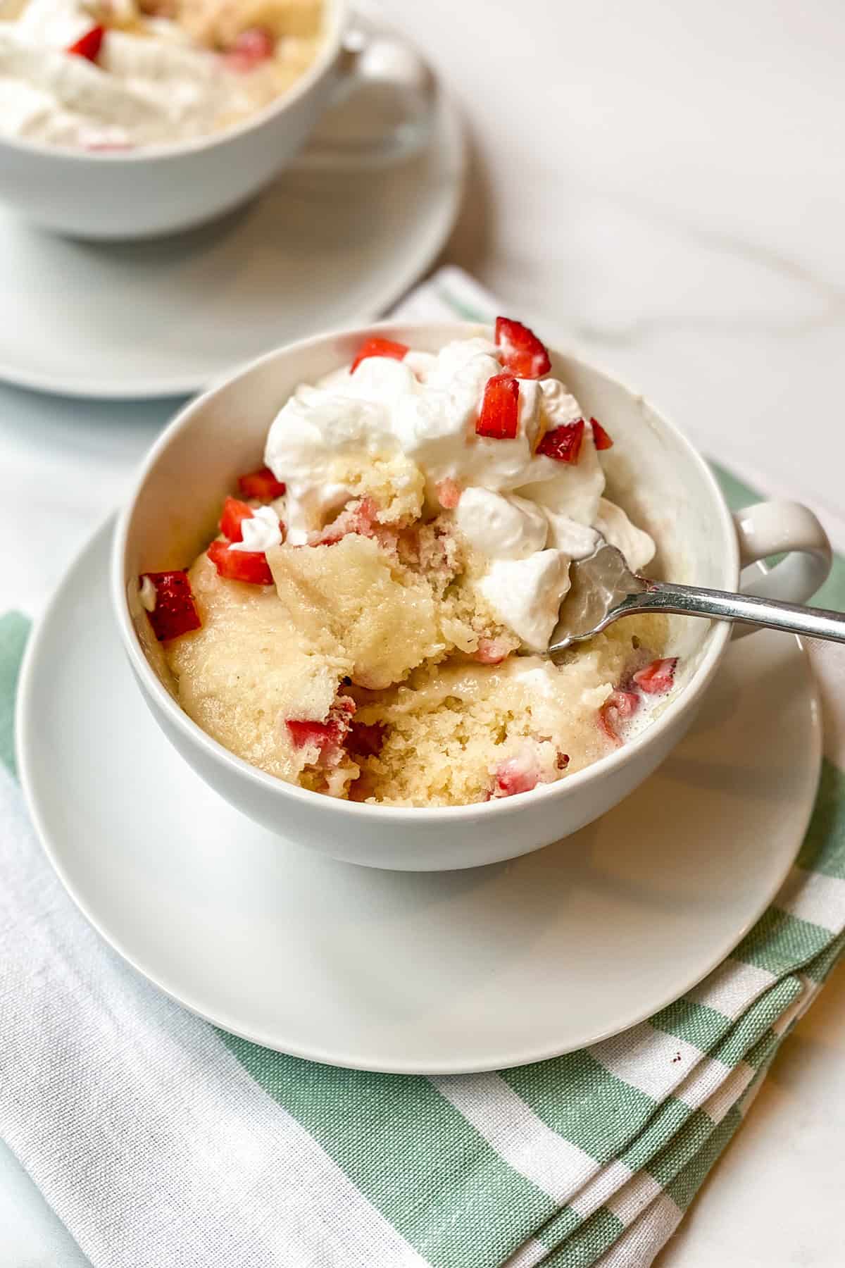 A cup of strawberry mug cake topped with whipped cream and strawberry pieces, with a spoonful taken out, sits on a saucer over a folded napkin.