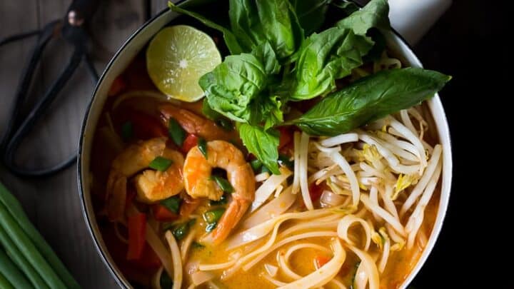A pot of shrimp noodle soup garnished with fresh basil, bean sprouts, and lime—surrounded by green onions, basil leaves, and ginger—makes for one of the best healthy 20 minute dinners on a wooden table.