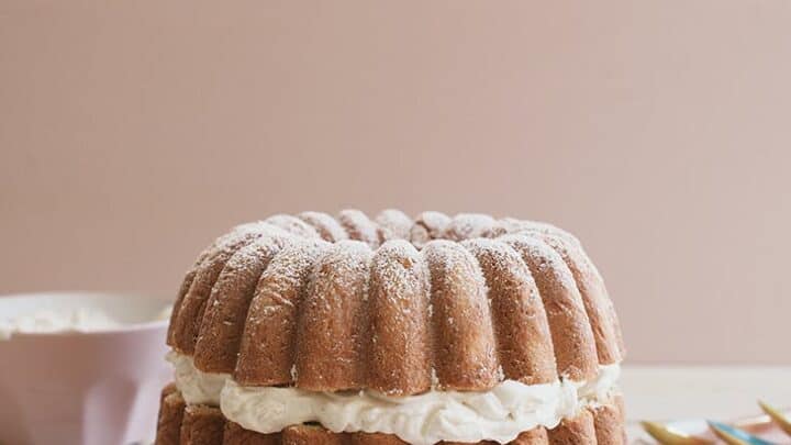 A round bundt cake with a layer of whipped cream filling, dusted with powdered sugar, sits on a plate next to utensils and a bowl—perfect for enjoying during fika.