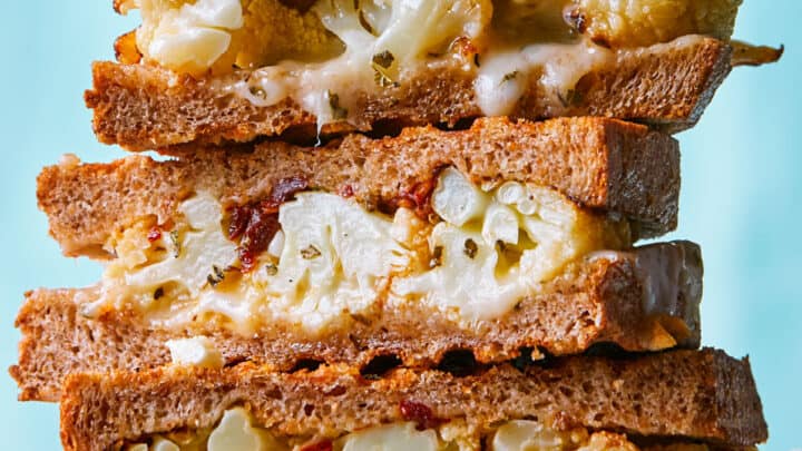 A stack of three grilled cheese sandwiches filled with roasted cauliflower, melted cheese, and bits of sun-dried tomato on whole wheat bread—perfect winter comfort food—set against a light blue background.