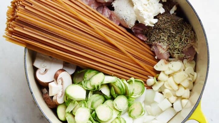 Yellow pot containing uncooked spaghetti, raw chicken, sliced mushrooms, Brussels sprouts, chopped onion, garlic, ricotta cheese, and dried herbs—perfect for cozy January One Pot Dinners on a white surface.