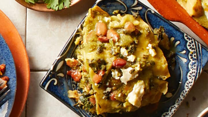 Square plate with green enchiladas topped with cheese and beans, surrounded by a bowl of cilantro and other dishes on a tiled surface—perfect winter comfort food for cozy nights.