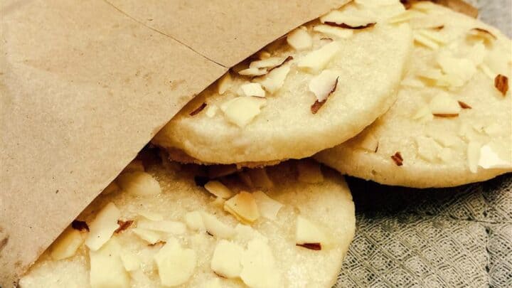 Three almond cookies, perfect for fika, partially inside a brown paper bag, resting on a textured gray surface.