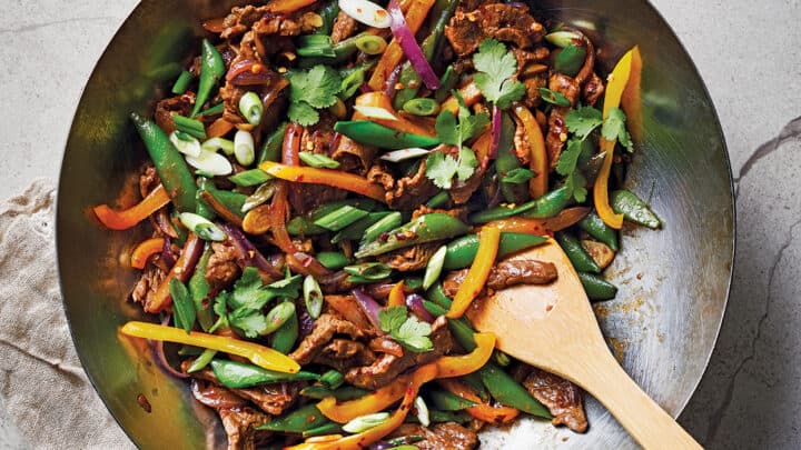 A wok filled with stir-fried beef, bell peppers, snap peas, red onions, and herbs makes for one of the perfect 400 Calorie Dinners, with a wooden spatula resting inside.