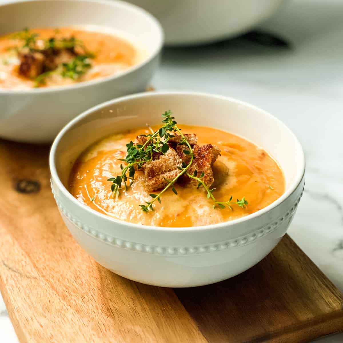 Square image of creamy root vegetable soup in a white bowl garnished with fresh thyme and toasted croutons.