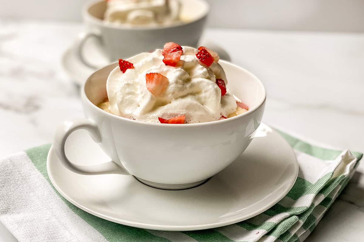 A white cup filled with strawberry mug cake, topped with whipped cream and strawberry pieces, sits on a saucer, with another cup in the background.