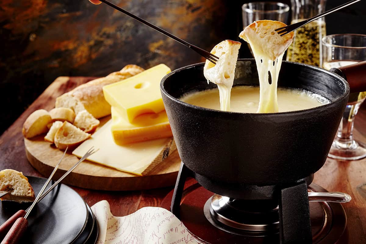 Slices of bread are dipped into creamy fondue in a pot, with cheese, bread, and wine glasses artfully arranged on a rustic wooden table.