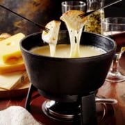 Two forks hold bread dipped in creamy fondue in a black pot, with Swiss cheese blocks and glasses in the background.