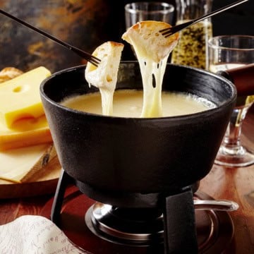 Two forks hold bread dipped in creamy fondue in a black pot, with Swiss cheese blocks and glasses in the background.