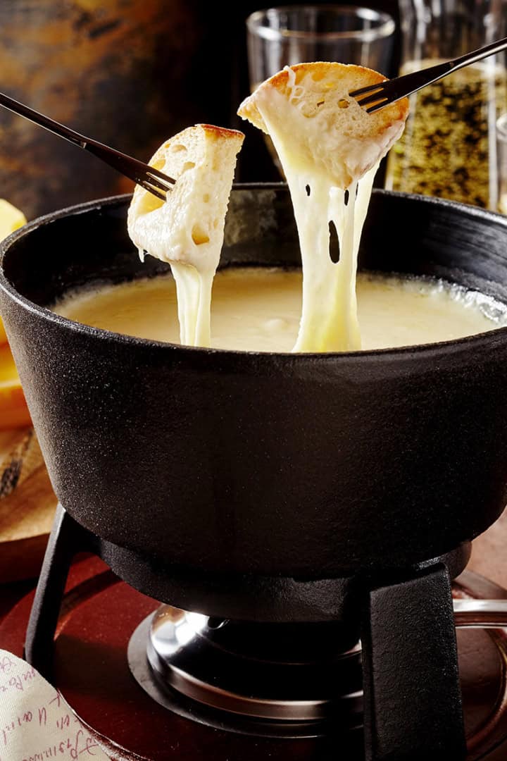 Two forks hold bread dipped in creamy fondue in a black pot, with Swiss cheese blocks and glasses in the background.