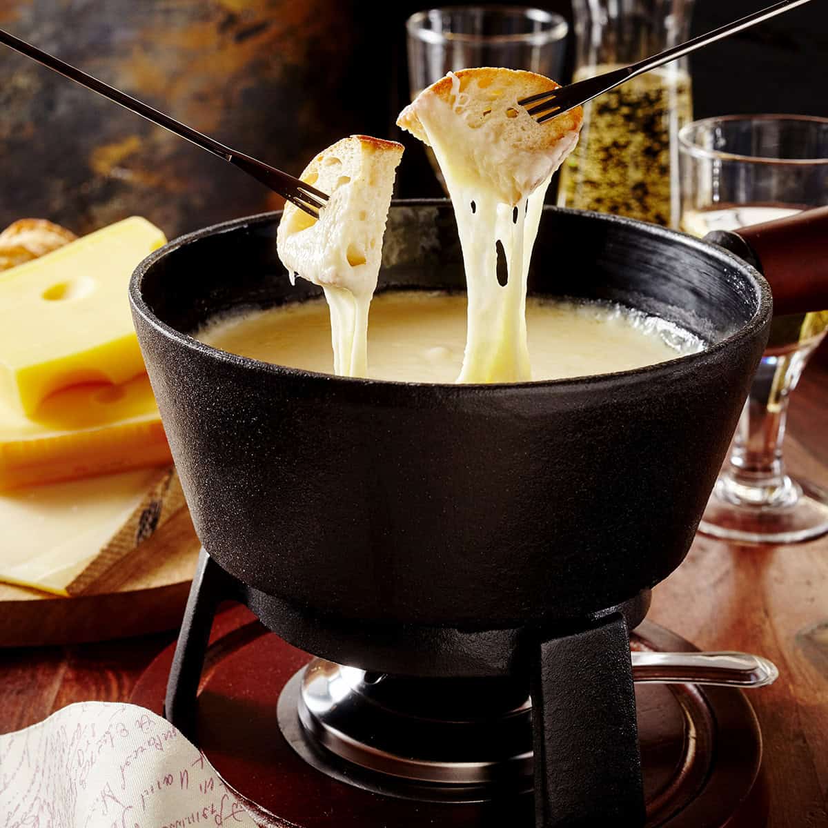 Two forks hold bread dipped in creamy fondue in a black pot, with Swiss cheese blocks and glasses in the background.